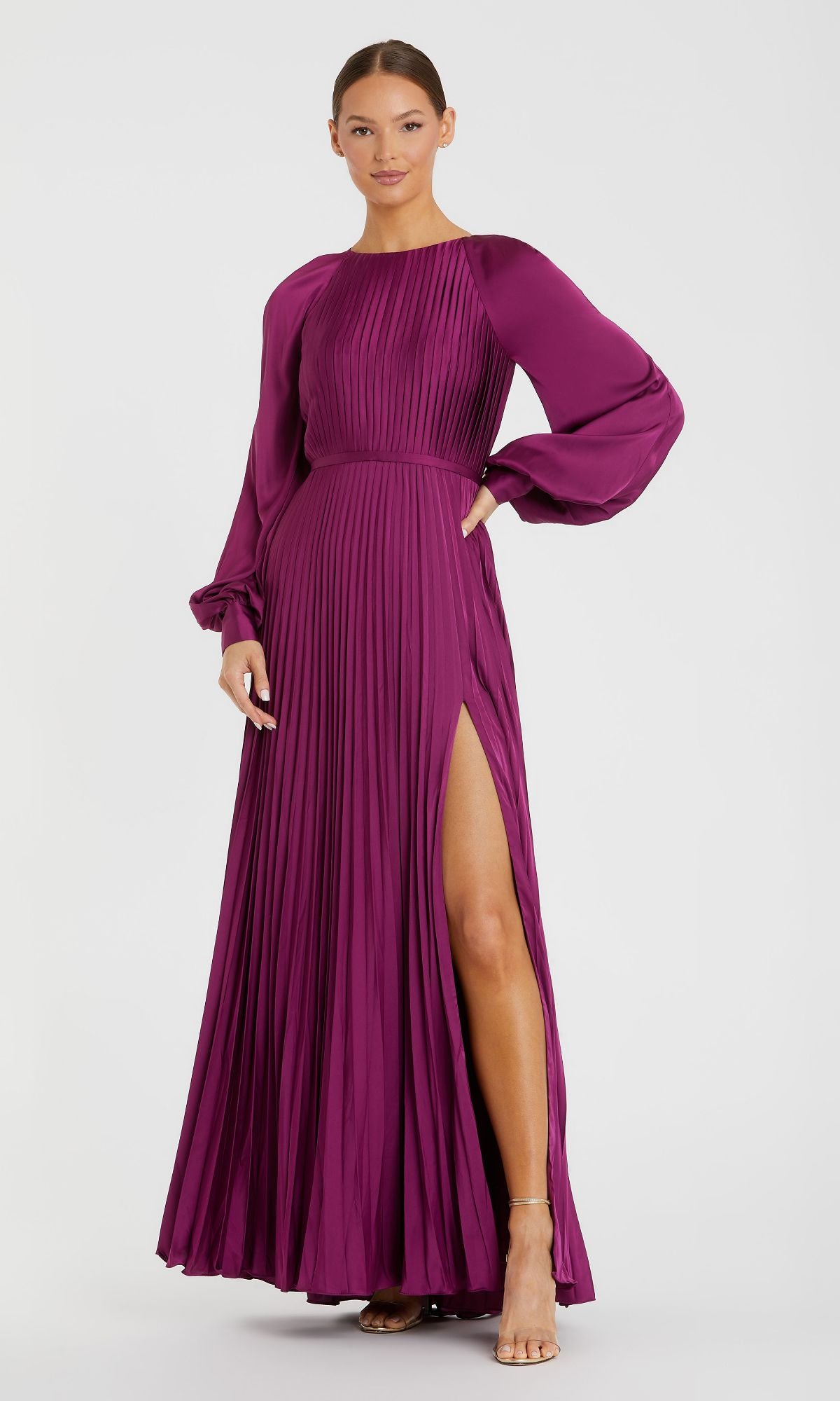 A woman wears the Mac Duggal 26590, a long sleeve pleated formal dress with a high slit, posing against a plain white background.